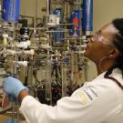 A researcher in a lab coat and safety glasses operates a complex setup of lab equipment.
