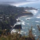 Coastal cliffs lined with houses overlooking the ocean, with rugged rock formations and waves crashing below.